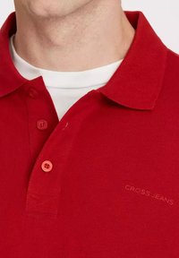 Czerwona koszulka polo z teksturowanej dzianiny, z klasycznym kołnierzykiem, trzema guzikami oraz subtelnym logo "CROSS JEANS" z boku.