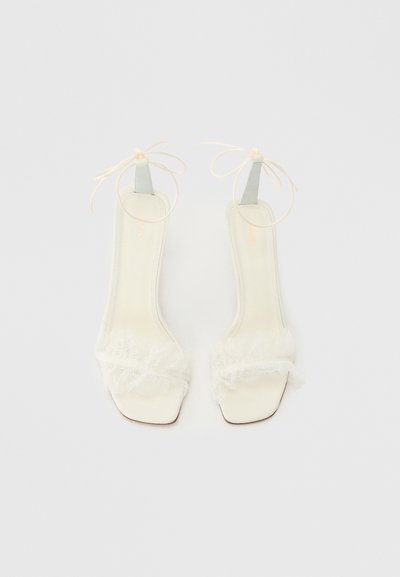 Λευκά σανδάλια ανοιχτά μπροστά με λεπτά λουράκια στον αστράγαλο δεμένα σε φιόγκους και λεπτή δαντέλα πάνω από τα δάχτυλα, εμφανιζόμενα από πάνω σε απλό φόντο.