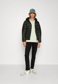 Schwarze, puffy Jacke, leicht, mit Reißverschluss und Kapuze. Hellgrünes T-Shirt darunter, schwarze Hose und weißes Sneakers mit grünen Akzenten.
