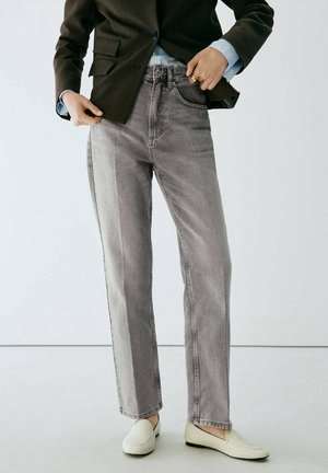 Personne portant un jean droit gris bicolore, un blazer noir, une chemise bleu clair et des chaussures blanches à enfiler, se tenant devant un fond blanc uni.