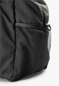 Primer plano de una mochila negra que muestra un bolsillo lateral de malla y un lazo de plástico cerca de una cremallera en la sección superior.