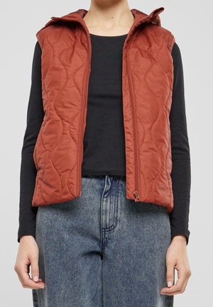 Personne portant un gilet matelassé sans manches couleur rouille sur un t-shirt noir à manches longues et un jean bleu, debout devant un fond uni.