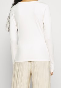 Modella con un top bianco a maniche lunghe a coste abbinato a pantaloni beigi pieghettati, che mostra la vista posteriore e la vestibilità rilassata dell'outfit.