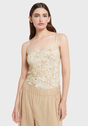Jeune femme aux longs cheveux bruns portant un haut beige à fines bretelles avec imprimé floral et un pantalon beige ample, sur fond uni.