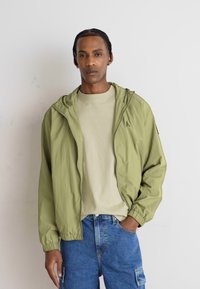 Giacca a zip verde oliva chiaro con cappuccio e polsini elasticizzati, indossata sopra una t-shirt beige, abbinata a jeans in denim blu.