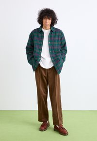 Zöld és tengerészkék kockás flanel ing, alatta fehér póló, barna selyemhatású nadrág és bordó fűzős cipő. Egyszínű háttér.