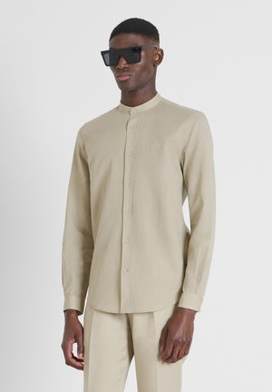 Beige overhemd met lange mouwen en een Mandarin-kraag, knoopsluiting en subtiele logo. Draagt zwarte zonnebril voor een effen achtergrond.