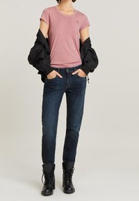 Person trägt ein pinkes T-Shirt, dunkelblaue Jeans, schwarze Schnürstiefel und eine schwarze Jacke, die über die Schultern drapiert ist.