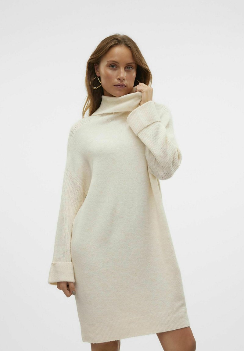 Abito maglione lavorato a maglia, oversize, di colore crema con colletto alto e polsini a costine. Presenta maniche testurizzate e una vestibilità ampia e rilassata.