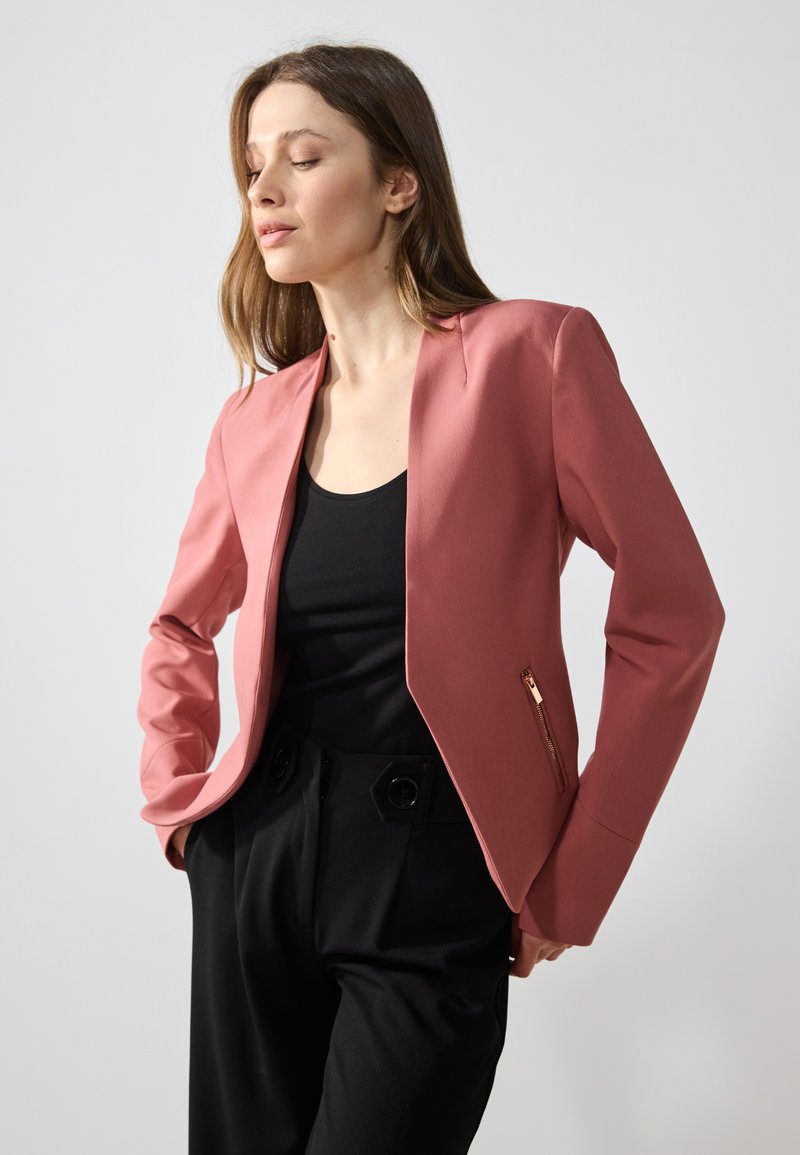 Roze op maat gemaakte blazer met een gladde textuur, ritszakdetails en lange mouwen, gecombineerd met een zwarte top en hooggetailleerde broek.