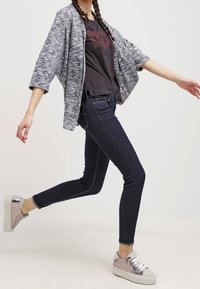 Persona vistiendo un cárdigan gris abierto, camiseta negra, jeans oscuros ajustados y zapatillas plateadas, posando en medio de un paso contra un fondo liso.