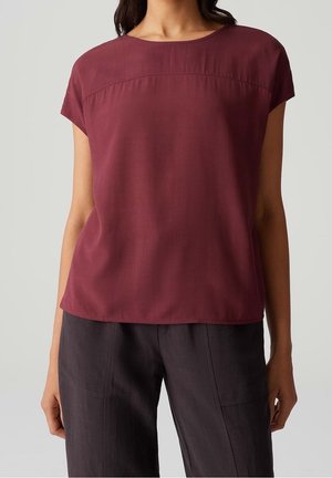 Femme portant un chemisier bordeaux à manches courtes associé à un pantalon ample gris foncé, debout devant un fond clair uni.