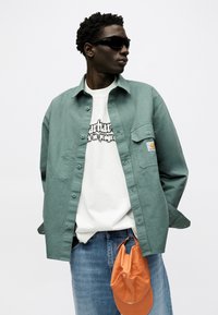 Muž s černými slunečními brýlemi, zelenou košilí na knoflíky přes bílý tričko s potiskem, modré džíny, drží v ruce oranžovou baseballovou čepici, dívá se doprava.