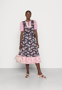 Vestido floral com mangas bufantes cor-de-rosa, base azul-marinho, decorado com flores. Apresenta uma bainha com volantes e uma pequena bolsa cor-de-rosa.