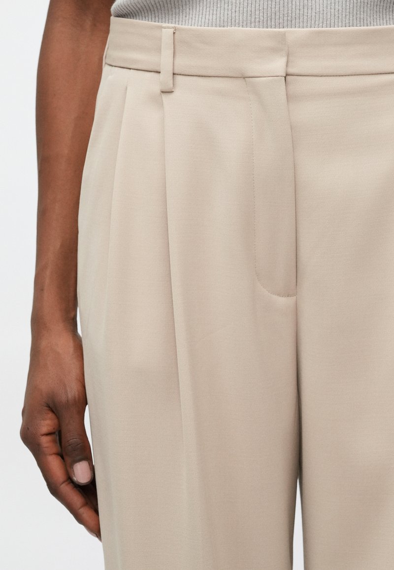 Pantalones beige de tejido suave, con pliegues en la parte delantera, bolsillo lateral y un corte entallado con cintura plana.