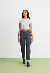 Grijze cropped t-shirt, zwarte recht gesneden broek en zwarte sneakers met witte accenten. Model heeft lange vlechten en staat op een groene vloer.