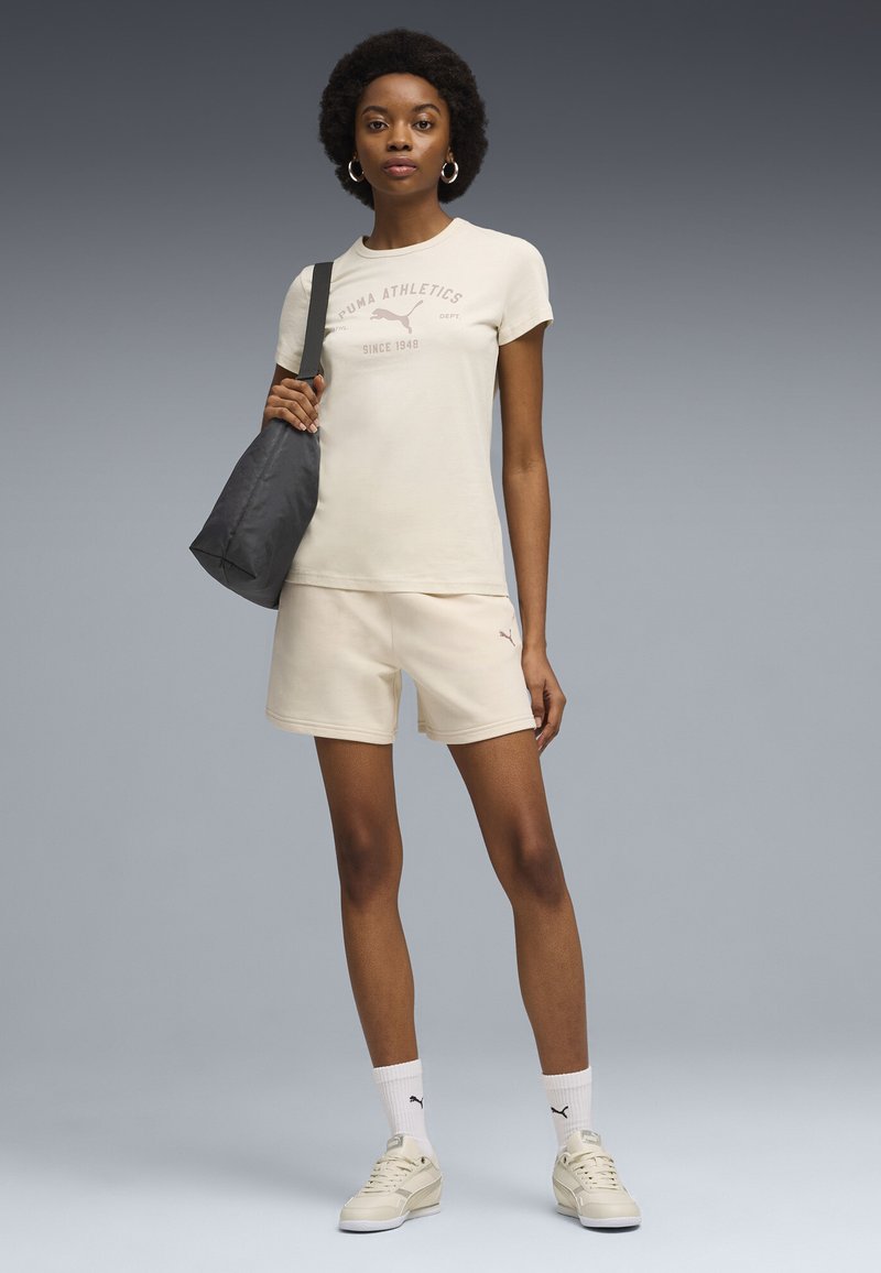 Kobieta w beżowej koszulce i szortach Puma, białych skarpetach Puma, kremowych trampkach, trzymająca czarną torbę na ramię, stojąca na tle szarego tła.