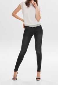 Blusa blanca de manga corta con textura de puntos, combinada con jeans ajustados negros. La modelo lleva sandalias de tacón negro.