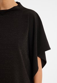 Schwarzes, kurzärmeliges Top mit hohem Kragen und einer weichen, strukturierten Oberfläche, die dezent metallische Punkte aufweist.