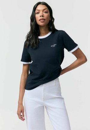 Kobieta o długich ciemnych włosach, ubrana w granatowy T-shirt z białą lamówką i białe spodnie, stojąca z jedną ręką na biodrze.