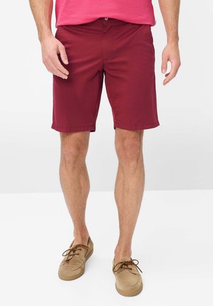 Mann trägt burgunderrote knielange Shorts, ein rosa Hemd und beige Schnür-Mokassins, steht vor einem schlichten weißen Hintergrund.