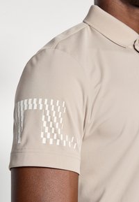 Beige polo skjorte laget av glatt stoff, med krage og korte ermer. Hvite mønstrede detaljer på venstre erme gir et ekstra preg.