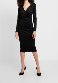 Schwarzes langärmliges Kleid aus weichem Stoff, mit V-Ausschnitt und gerüschten Details, knielanger Schnitt, kombiniert mit schwarzen hohen Schuhen.