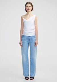 Witte top met hoge kraag, gecombineerd met lichtblauwe rechte jeans met verspreide witte embellissementen; model staat op zwarte open teen hakken.