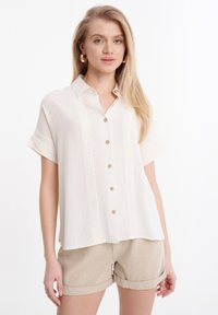 Camicia beige a maniche corte con bottoni, dotata di dettagli in pizzo sul davanti, caratterizzata da una vestibilità rilassata e un tessuto morbido e testurizzato. Abbinata a pantaloni corti beige.