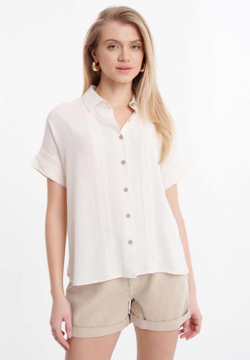 Beige kortemouwen blouse met kantdetails aan de voorkant, met een losse pasvorm en zachte, textuurrijke stof. Gepaard met beige shorts.