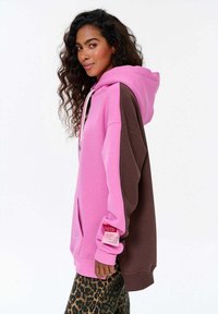 Nadměrná mikina v růžové a hnědé barvě, s kapucí na stažení, přední kapsou a textilním štítkem na rukávu. Měkký materiál z bavlněné směsi.