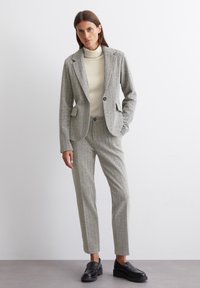 Grau strukturierte Blazerjacke mit passenden taillierten Hosen, ergänzt durch ein cremefarbenes Oberteil mit hohem Kragen und schwarzen Slippers. Strukturiertes Design mit Taschen.