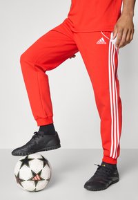 Røde treningsbukser laget av glatt stoff, med tre hvite striper langs sidene, sammen med svarte fotballsko på en mønstret ball.