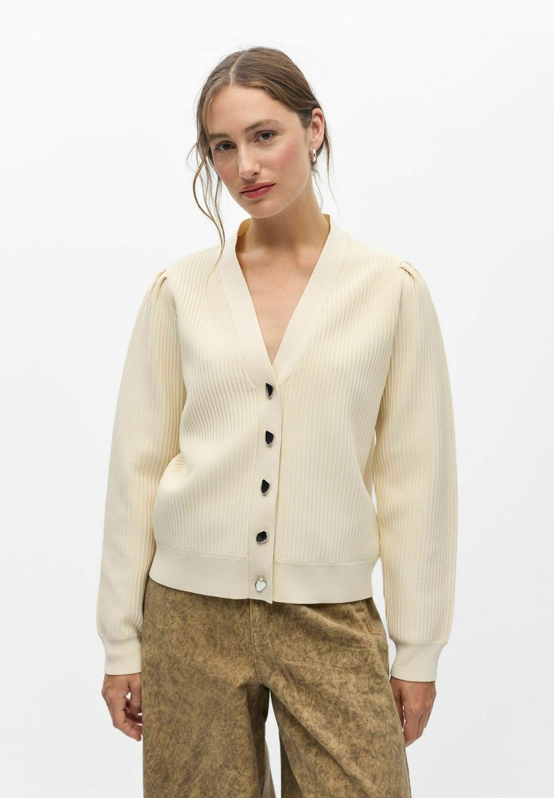 Femme portant un cardigan côtelé crème avec des boutons noirs et un pantalon marron texturé, debout sur un fond blanc.