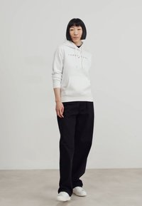Felpa bianca con cappuccio e tasca frontale, caratterizzata da un logo; abbinata a pantaloni neri a gamba larga e sneakers bianche. Texture morbida, design casual.
