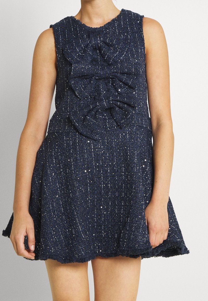 Robe sans manches bleu foncé texturée avec des taches blanches et trois grands nœuds en tissu sur le devant, portée par une personne debout.