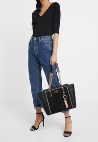 Mustassa tekstuurilla varustettu olkalaukku, jossa on kultaketju ja vaaleanpunaiset yksityiskohdat. Laukussa on avaimenperä ja suorakulmainen muoto, ja se on yhdistetty mustan paidan ja sinisten farkkujen kanssa.