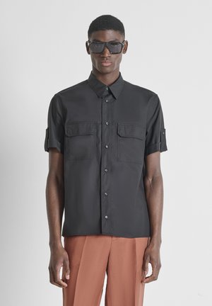 Man draagt een zwart overhemd met korte mouwen en knopen met voorzakken, bruine broek en donkere zonnebril, staand tegen een effen achtergrond.