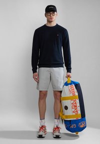 Marineblå sweater, lysegrå shorts og taske i flere farver. Tilbehøret inkluderer en sort kasket og briller. Skoene er hvide med orange detaljer.
