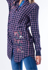 Camicia a quadretti blu navy e rosa con maniche lunghe, caratterizzata da ricami floreali su un lato e orlo curvo. Realizzata in tessuto leggero.