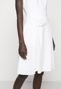 Bílé šaty s přiléhavým korzetem a detaily ovinutého pasu. Hladká tkanina s lehce splývavými sukněmi a bočním rozparkem.