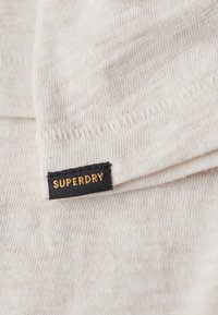 Tessuto in cotone beige chiaro con una texture a righe, con un'etichetta nera con "SUPERDRY" in lettere dorate.
