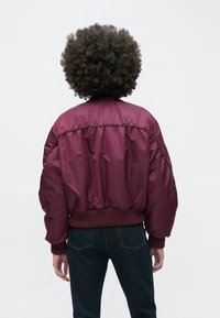 Sarkanvīna krāsas bomber jaka ar spīdīgu audumu, adītām aprocēm un apakšmalu, kā arī pārmērīgu siluetu, skatīta no aizmugures pret gaišu fonu.