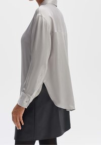 Blouse gris clair avec un col, des manches longues, un ourlet arrondi et des poignets boutonnés, associée à une jupe noire. Texture de tissu lisse.