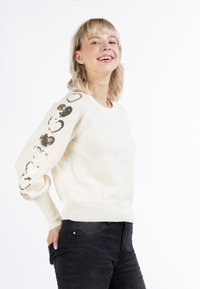 Krém színű pulóver hosszú ujjal, amelyen fémes arany szívalakú flitterek vannak az egyik ujj mentén. Bordás mandzsetták és alsó rész. Sötét farmernadrággal párosítva.