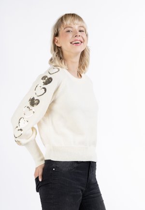 Pull crème avec des manches longues orné de sequins en forme de cœur dorés métalliques le long d'une manche. Poignets et ourlet côtelés. Assorti avec un jean foncé.