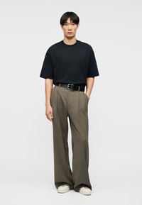 Άνδρας που στέκεται με τα χέρια στις τσέπες, φοράει μαύρο oversize t-shirt, καφέ παντελόνι με φαρδιά μπατζάκια και μαύρη ζώνη, καθώς και λευκά αθλητικά παπούτσια σε απλό φόντο.