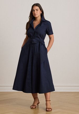Une femme pose dans une robe-chemise bleu marine à manches courtes, avec une jupe midi évasée et une taille nouée. Elle a les mains dans les poches.