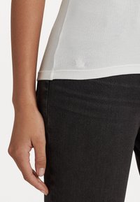 Bílý žebrovaný top s kulatým lemem, doplněný jemnými detaily šití, spárovaný s tmavými džíny. Ruka spočívá na bocích.