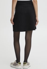 Jupe noire avec une finition lisse, ourlet tombant au milieu de la cuisse, associée à des collants noirs transparents et des chaussures de sport grises avec des accents verts.